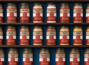 The word STAPLES is spelled out using different spices, herbs, and salt, with small sprigs of rosemary, garlic cloves, and scattered peppercorns on a dark surface.