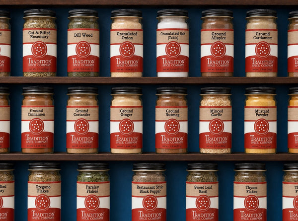 The word STAPLES is spelled out using different spices, herbs, and salt, with small sprigs of rosemary, garlic cloves, and scattered peppercorns on a dark surface.