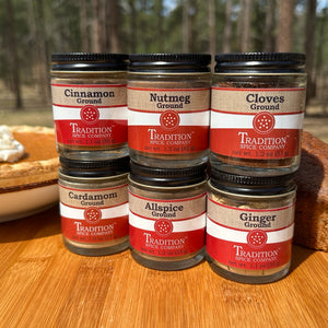 Six jars of ground spices from Tradition Spice Company on a wooden surface with a blurred natural background.