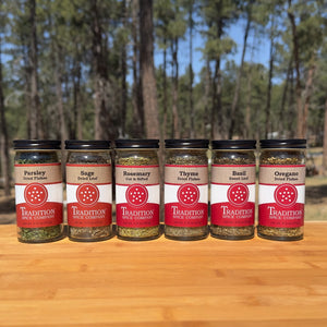 Six jars of parsley, sage, rosemary, thyme, basil, and oregano from Tradition Spice Company's Basic Herbs - Pantry Pack 2 are lined up on a wooden surface with a forest backdrop, creating the ultimate kitchen herbs bundle.