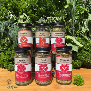 Six jars from Basic Herbs - Pantry Pack 2—parsley, sage, rosemary, thyme, basil, and oregano—are stacked in two rows on a wooden surface with fresh herbs and greenery behind. Ideal pantry essentials for any kitchen.