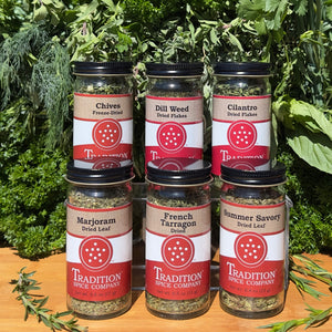 Six jars of Basic Spices - Pantry Pack 1—chives, dill weed, cilantro, marjoram, French tarragon, and summer savory—are arranged in two rows as a perfect dried herbs bundle for pantry staples. Fresh greenery decorates the background.