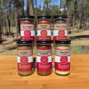 Basic Spices - Pantry Pack 2 from Tradition Spice Company includes red pepper, cayenne, black pepper, cumin, smoked sweet Spanish paprika, and mustard powder displayed on wood with a forest backdrop.