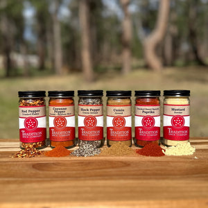 Six jars of Basic Spices - Pantry Pack 2—red pepper, cayenne, black pepper, cumin, paprika, and mustard powder—are displayed on a wooden surface outdoors with a blurred natural background.