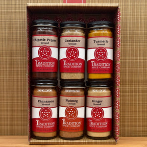 The Soup Toppers Savory Set includes six Tradition Spice Company jars—chipotle pepper, coriander, turmeric, cinnamon, nutmeg, and ginger—neatly boxed in two rows of three against a brown plaid background.