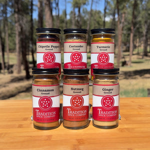 Six glass jars from the Soup Toppers Savory Set—labeled Chipotle Pepper, Coriander, Turmeric, Cinnamon, Nutmeg, and Ginger—are arranged on a wooden surface with a forest backdrop.
