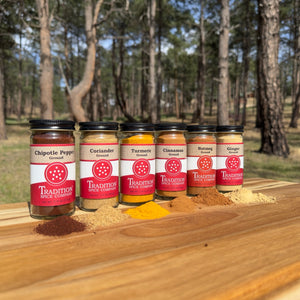 The Soup Toppers Savory Set features six ground spices—Chipotle Pepper, Coriander, Turmeric, Cinnamon, Nutmeg, and Ginger—displayed outdoors on a wooden surface with pine trees in the background and spice piles in front.