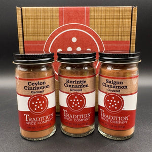 Three glass jars of ground spices from Tradition Spice Company, including Ceylon Cinnamon, Korintje Cinnamon, and Saigon Cinnamon, arranged in front of their branded cardboard gift box on a dark background.