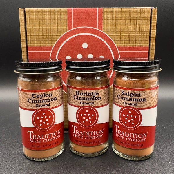 Three glass jars of ground spices from Tradition Spice Company, including Ceylon Cinnamon, Korintje Cinnamon, and Saigon Cinnamon, arranged in front of their branded cardboard gift box on a dark background.