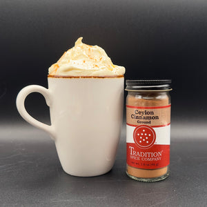 A white ceramic mug filled with a whipped hot drink and a sprinkle of cinnamon, standing next to a glass jar of Tradition Spice Company Ceylon Cinnamon Ground on a dark gray background.