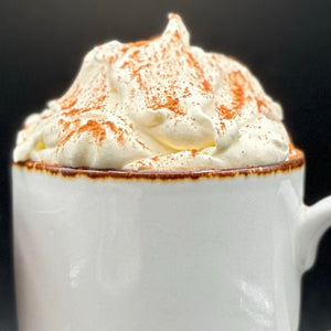 A close-up of a white ceramic mug filled with a hot beverage topped with thick whipped cream and dusted with cinnamon, set against a dark background.