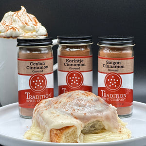 Three jars of ground cinnamon from Tradition Spice Company are displayed behind a cinnamon roll topped with cream and cocoa powder, with a whipped drink in the background on a dark studio backdrop.