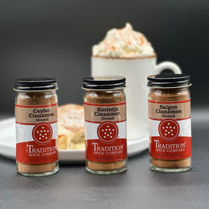 Three glass spice jars labeled Ceylon Cinnamon, Korintje Cinnamon, and Saigon Cinnamon from Tradition Spice Company, arranged on a dark surface with a blurred coffee cup in the background.