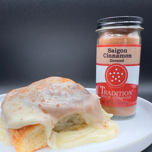 A jar of Tradition Spice Company Saigon Cinnamon Ground next to a cinnamon roll with white icing on a white plate, set against a dark gray background.