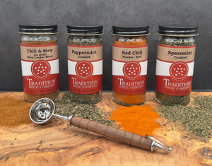 Four jars of spice blends from Tradition Spice Company are arranged on a wooden surface with scattered spices and a metal measuring spoon in front. Spices include chili, peppermint, red chili, and spearmint.