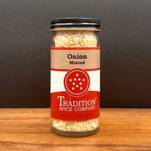 A glass jar of Tradition Spice Company Onion Minced spice blend, featuring a black metal lid and a red and white label, set on a wooden surface against a dark gray background.