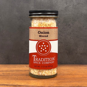 A glass jar of Tradition Spice Company Onion Minced spice blend, featuring a black lid and a red and white label, set on a wooden surface against a dark gray background.