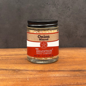 A glass jar of Tradition Spice Company Onion Minced seasoning with a black lid, set on a wooden surface against a dark gray background.