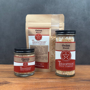 Three jars and one large bag of onion mince spice from Tradition Spice Company arranged on a wooden surface against a dark gray background.
