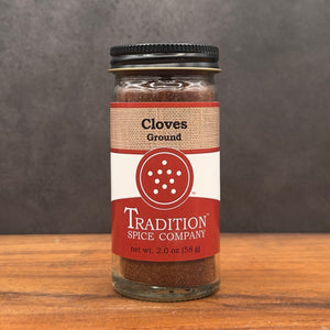 A glass jar of Ground Cloves with a black lid and brown-red label sits on a wooden surface against a gray background—an aromatic spice perfect for enhancing both sweet and savory dishes.