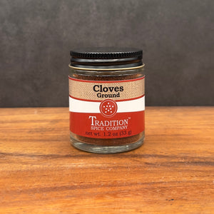 A 1.2 oz (33 g) glass jar of Ground Cloves from Tradition Spice Company sits on wood, featuring a red and white label—ideal for sweet or savory recipes.