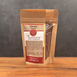A brown paper pouch of Ground Cloves stands on a wooden surface, featuring the product name, description, and ingredients on a red and cream label. This aromatic spice is ideal for enhancing both sweet and savory dishes.