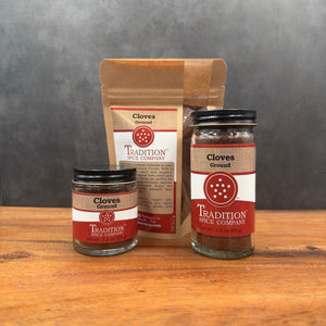 Three containers of Tradition Spice Company’s Ground Cloves—a small glass jar, medium glass jar, and brown resealable pouch with red and white labels—are displayed on a wooden surface, perfect for sweet and savory dishes.