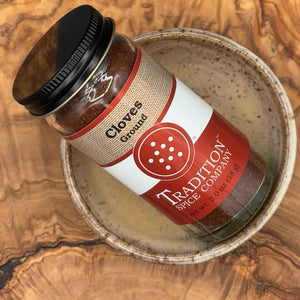 A jar of Ground Cloves sits in a ceramic bowl on a wooden surface with visible natural grain, ready to enhance sweet or savory dishes.