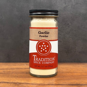 A glass jar of Tradition Spice Company Garlic Powder on a wooden surface against a dark gray background.