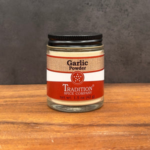 A glass jar of Tradition Spice Company Garlic Powder with a black lid, set on a wooden surface against a dark gray background.