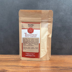 A stand-up kraft paper pouch of Tradition Spice Company Garlic Powder, standing on a wooden surface against a dark gray textured background.