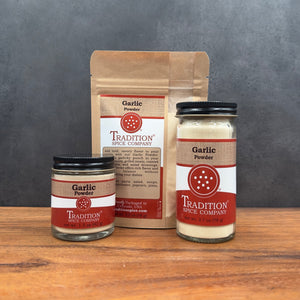 Three jars and one stand-up pouch of garlic powder from Tradition Spice Company arranged on a wooden surface against a dark gray backdrop.