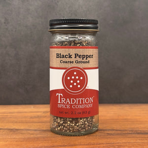 A glass jar labeled Course Ground Black Pepper from Tradition Spice Company sits on a wooden surface with a gray background, delivering bold, rustic seasoning in every sprinkle. The jar contains coarse ground black pepper.