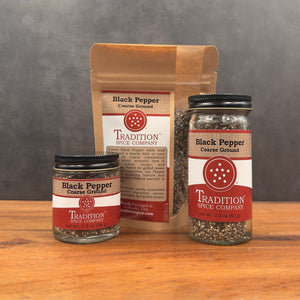 Three containers of Course Ground Black Pepper—two glass jars and one brown paper pouch—are displayed on wood, highlighting this bold seasoning against a gray backdrop.