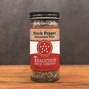A glass jar of Restaurant Style Black Pepper with a black lid rests on a wooden surface against a dark background. The tan and red label displays white and black text, highlighting its bold seasoning flavor.