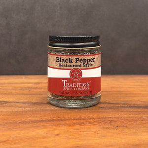 A glass jar of Restaurant Style Black Pepper with a bold flavor sits on a wooden surface against a dark backdrop. The white and red label identifies the 1.3 oz (37g) jar, featuring a sleek black lid for freshness.