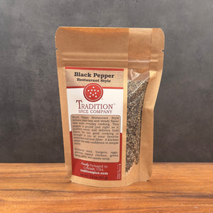 A brown resealable pouch labeled "Restaurant Style Black Pepper" sits on a wooden surface with visible black pepper granules through a clear window, highlighting its bold seasoning against a gray background.