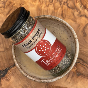 A jar of Restaurant Style Black Pepper lies on its side inside a speckled ceramic bowl, resting on a wooden surface.