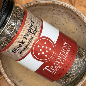 A jar of Restaurant Style Black Pepper rests at an angle in a ceramic bowl, its red and tan label featuring white text and a pepper shaker graphic suggesting the bold flavor inside.
