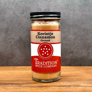 A glass jar of Tradition Spice Company Korintje Cinnamon Ground, showing the light brown ground cinnamon inside, set on a wooden surface against a textured gray background.