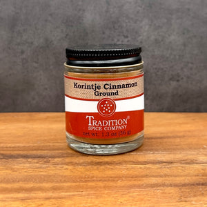 A glass jar labeled Ground Saigon Cinnamon from Tradition Spice Company sits on a wooden surface with a gray background. The jar, filled with premium cinnamon, features a black lid and an orange and white label—making it an excellent addition to your collection of baking spices.