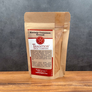 A brown resealable pouch of Tradition Spice Company Ground Saigon Cinnamon, a premium cinnamon prized among baking spices, sits on a wooden surface with a gray background. The front label describes its uses and highlights its sweet, warm flavor.