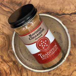 A jar of Ground Saigon Cinnamon from Tradition Spice Company sits on a ceramic dish atop a wooden surface, showcasing one of the finest baking spices for your kitchen.