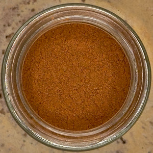 A top-down view of a glass jar filled with finely ground reddish-brown spice, identified as Ground Saigon Cinnamon, on a beige surface—perfect for adding warmth and depth to your favorite baking spices.