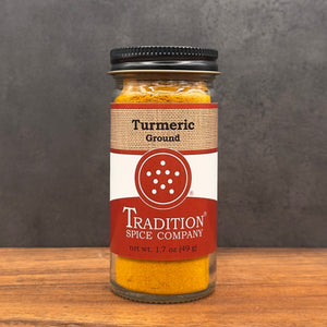 A glass jar of Tradition Spice Company Turmeric Ground spice, filled with bright yellow-orange powder and topped with a black lid, set on a wooden surface against a dark gray background.