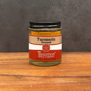 A jar of Tradition Spice Company Turmeric Ground spice on a wooden surface against a dark gray textured background.