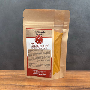 A stand-up kraft paper pouch of Tradition Spice Company Turmeric Ground, showing the yellow powder through a clear window on a wooden surface against a gray background.