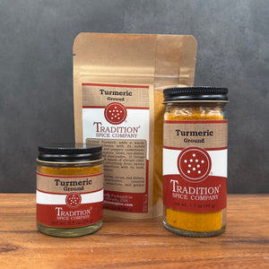 Three packages of Tradition Spice Company Turmeric Ground spice displayed on a wooden surface against a dark gray background: a large kraft paper pouch in the back, a small glass jar on the left, and a larger glass jar on the right.