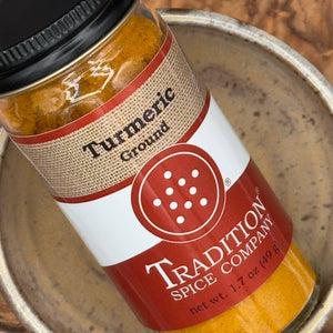 A close-up of a glass jar of Tradition Spice Company ground turmeric spice, showing the orange-yellow powder and label, resting inside a rustic ceramic bowl.