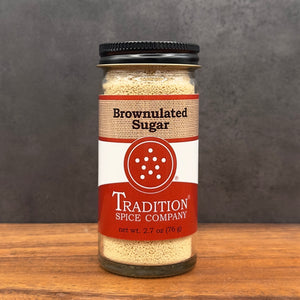 A glass jar of Tradition Spice Company Brownnulated Sugar with a black lid, set on a wooden surface against a dark gray background.